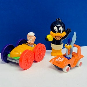 May include: A set of three vintage Looney Tunes toy figures. The figures are of Bugs Bunny, Daffy Duck, and Porky Pig. Bugs Bunny is driving a yellow car with orange wheels. Daffy Duck is standing with a yellow object in his hand. Porky Pig is driving a orange car with purple wheels. The cars have the words "Beep Beep!" printed on them.