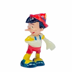 May include: A small plastic figurine of Pinocchio with a red hat, yellow shirt, red shorts, and blue shoes. His nose is long and pointed.