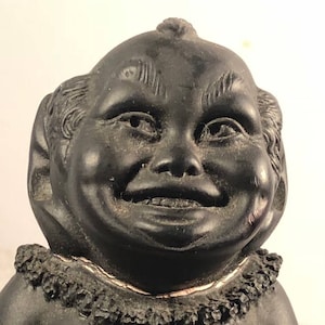 Op de afbeelding: Zwarte keramische figuur van een lachende, mollige persoon met een brede glimlach en een klein plukje haar op zijn hoofd. De figuur draagt een getextureerde, gerimpelde kraag.