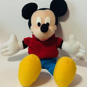 K&ouml;nnte beinhalten: Ein Pl&uuml;sch-Mickey-Mouse-Spielzeug, das ein rotes Hemd und eine blaue Hose tr&auml;gt. Das Spielzeug hat schwarze Ohren, eine schwarze Nase und gelbe Schuhe.