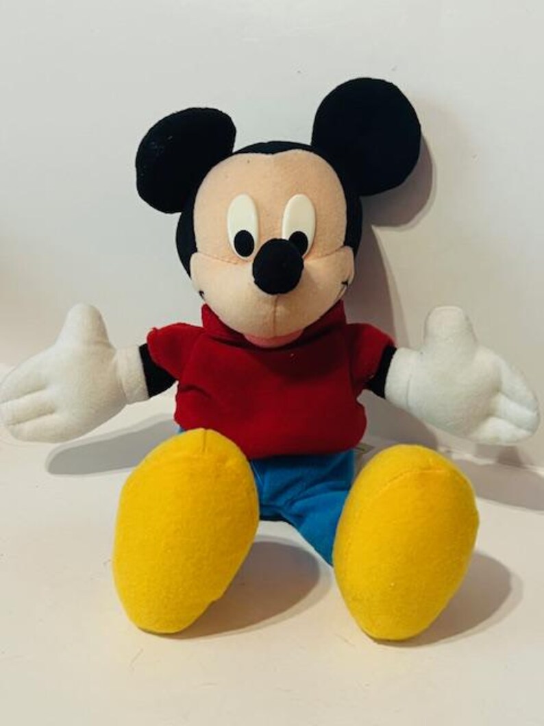 K&ouml;nnte beinhalten: Ein Pl&uuml;sch-Mickey-Mouse-Spielzeug, das ein rotes Hemd und eine blaue Hose tr&auml;gt. Das Spielzeug hat schwarze Ohren, eine schwarze Nase und gelbe Schuhe.