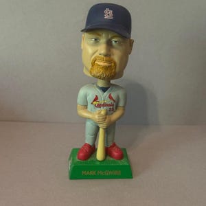 Könnte beinhalten: Eine Mark McGwire Wackelkopf-Figur. Die Figur trägt eine marineblaue Baseballkappe mit rotem Logo, ein graues Trikot mit dem Cardinals-Logo, rote Schuhe und hält einen Baseballschläger. Der Sockel ist grün mit dem Namen "MARK MCGWIRE".