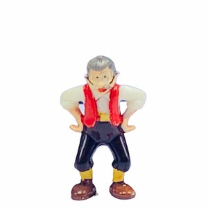 May include: A small figurine of a man with grey hair, a red waistcoat, black trousers, and brown shoes. He is standing with his hands on his hips and has a surprised expression on his face.
