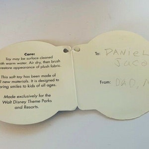 May include: White card with a care label for a plush toy. The care label includes instructions for cleaning and information about the toy's materials. The card also has a gift tag with the text "To: Daniel Jacobs" and "From: Dad, Mom".