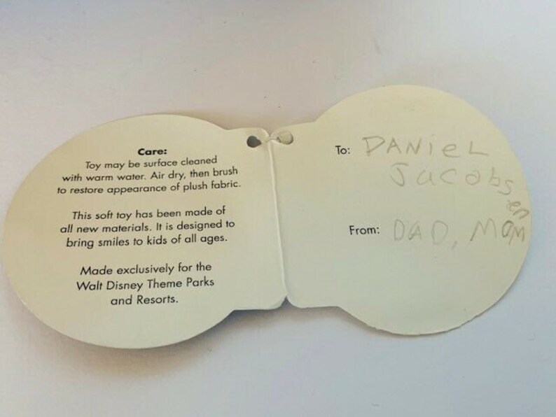 May include: White card with a care label for a plush toy. The care label includes instructions for cleaning and information about the toy's materials. The card also has a gift tag with the text "To: Daniel Jacobs" and "From: Dad, Mom".