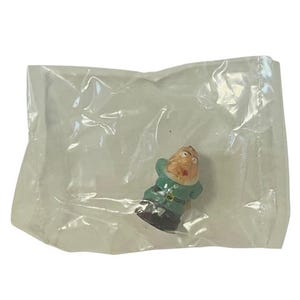 May include: A small plastic figurine of a man wearing a green and white outfit. The figurine has a surprised expression on its face.