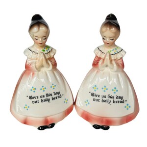 Pode incluir: Um par de saleiros e pimenteiros vintage com estatuetas de porcelana de meninas em vestidos cor-de-rosa com detalhes florais azuis. As meninas estão rezando com as mãos juntas. A base de cada saleiro tem o texto "Give us this day our daily bread" em letras pretas.