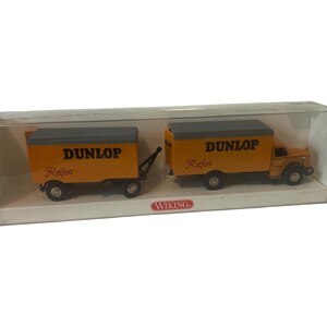 以下が含まれることがあります： グレーの屋根と側面に「DUNLOP」と書かれた黄色の玩具トラック。トラックは、グレーの屋根と側面に「DUNLOP」と書かれた黄色の玩具トレーラーを牽引しています。トラックとトレーラーはどちらも透明なプラスチック製のディスプレイケースに入っています。