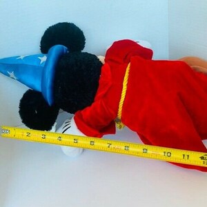 May include: A plush Mickey Mouse toy dressed as a magician. He is wearing a blue hat with white stars, a red robe, and a yellow rope around his waist. He is lying on a white surface with a measuring tape beside him.