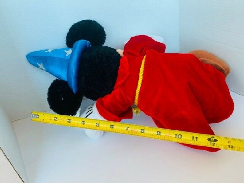 May include: A plush Mickey Mouse toy dressed as a magician. He is wearing a blue hat with white stars, a red robe, and a yellow rope around his waist. He is lying on a white surface with a measuring tape beside him.