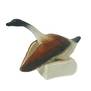 May include: A ceramic figurine of a goose with brown and white feathers, standing on a small white book. The goose has a black head and neck.