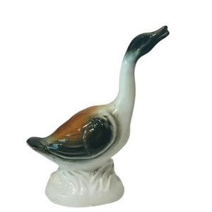 Puede incluir: Una figurita de ganso de cerámica pintada en marrón, negro y blanco. El ganso tiene la cabeza inclinada hacia arriba y el cuello curvado. La figurita está de pie sobre una base blanca.