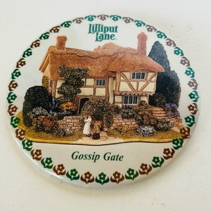 May include: A round ceramic plate with a green, brown, and white floral border. The center of the plate features a colorful illustration of a cottage with a thatched roof and the text "Lilliput Lane" and "Gossip Gate".