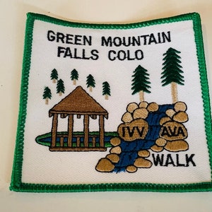 Pode incluir: Etiqueta bordada com fundo branco, borda verde e um gazebo marrom com um riacho azul e pedras. O texto "IVY AVA WALK" é bordado em preto. A etiqueta diz "Green Mountain Falls Colo".