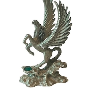 May include: A silver pewter figurine of a winged horse, or pegasus, with green gem accents. The pegasus is standing on a rocky base.