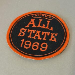 May include: A circular patch with an orange border and black center. The text "ALL STATE 1969" is in orange, with a crown design above the text. The patch is likely a vintage collectible or souvenir.