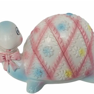May include: A ceramic turtle piggy bank with a pink and white quilted shell design. The turtle has a blue head and a pink bow tie.