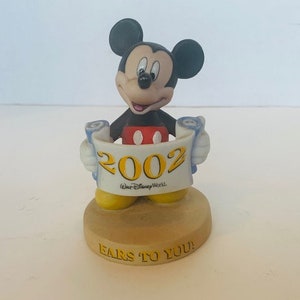 May include: A ceramic figurine of Mickey Mouse holding a scroll with the year 2002 and the text "Walt Disney World" on it. The base of the figurine is tan and has the text "Ears to You!" on it.