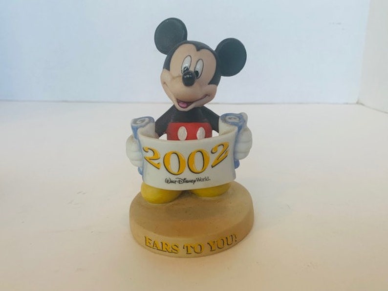 May include: A ceramic figurine of Mickey Mouse holding a scroll with the year 2002 and the text "Walt Disney World" on it. The base of the figurine is tan and has the text "Ears to You!" on it.