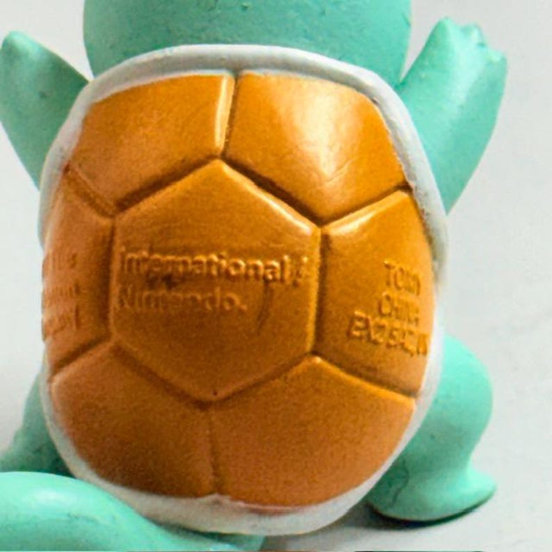 Puede incluir: Primer plano de una figura azul claro y naranja. La secci&oacute;n naranja tiene un patr&oacute;n geom&eacute;trico y el texto "International Nintendo". La figura es un personaje popular de un videojuego.