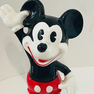 May include: A vintage ceramic figurine of Mickey Mouse. He is wearing his signature red shorts with white buttons and yellow shoes. Mickey is waving with his right hand and has a big smile on his face.