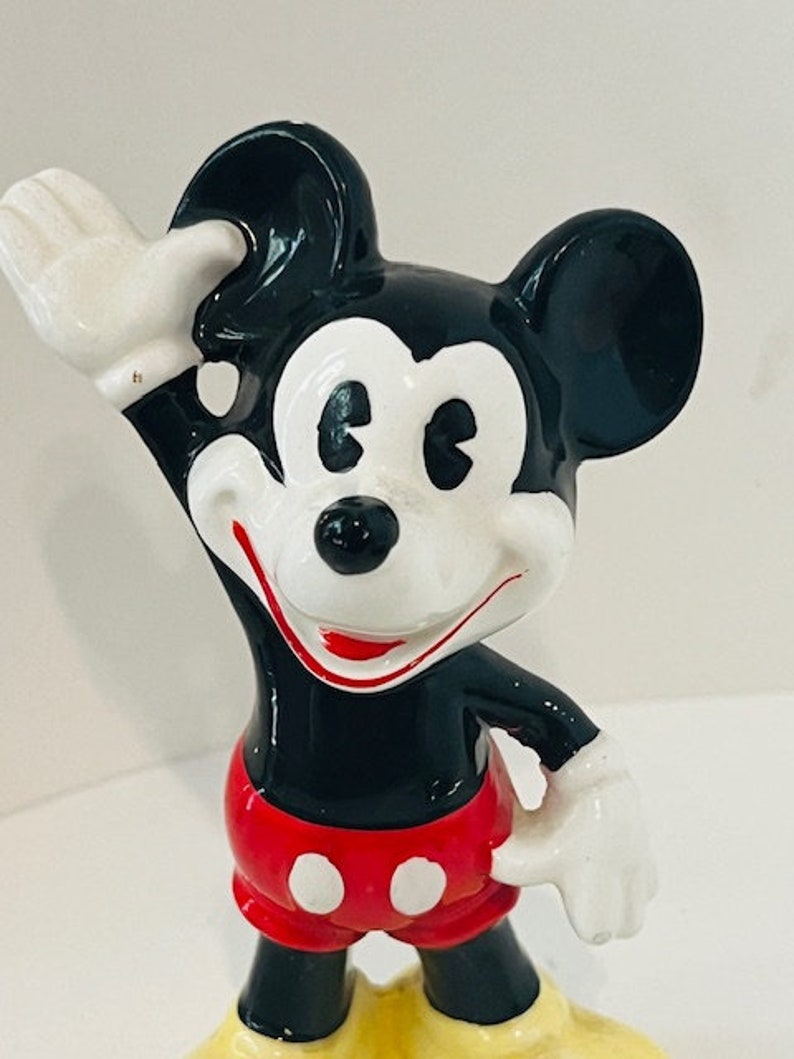 May include: A vintage ceramic figurine of Mickey Mouse. He is wearing his signature red shorts with white buttons and yellow shoes. Mickey is waving with his right hand and has a big smile on his face.