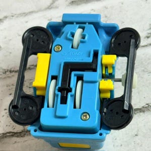 May include: A blue plastic toy vehicle chassis featuring four black wheels, two white wheels, and yellow accents. The underside is moulded with the text TOMY and SINGAPORE. The design includes a black central mechanism and metal axles for the wheels.