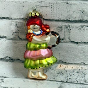 May include: A glass Christmas ornament shaped like a figure wearing a red hat, a blue necklace, and a layered dress with alternating lime green and light pink sections. The figure holds a round, dark-colored object and has a gold hanging loop.
