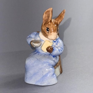 May include: A ceramic rabbit figurine in a blue dress, holding a small bowl and a yellow rectangle. The rabbit has brown fur and is seated on a small chair. The figurine has a classic, vintage aesthetic, reminiscent of a children's story.