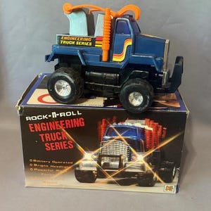 May include: A blue toy truck from the Rock-N-Roll Engineering Truck Series, with orange and yellow detailing. The truck is on a box with the series name and a truck image. The truck has large black tyres.