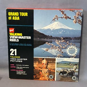 May include: A vintage "Grand Tour of Asia" View-Master reel set. The box displays a mountain and lake scene, alongside images of Asian culture. The text on the box reads "TALKING VIEW-MASTER REELS" and "21 TALKING VIEW-MASTER PICTURES".