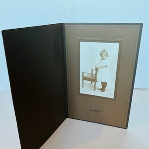 May include: A vintage photograph of a young girl in a white dress standing next to a small wooden chair. The photo is mounted in a brown cardstock frame with a black border.