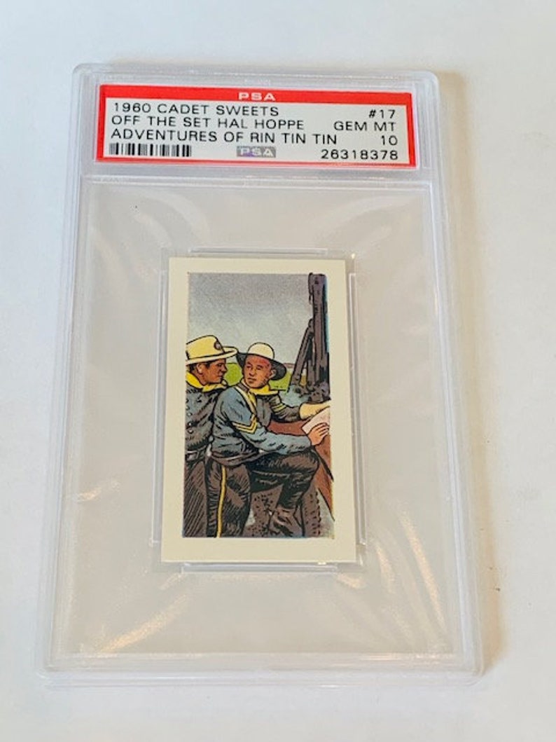 Pode incluir: Um cart&atilde;o classificado pela PSA que apresenta um cart&atilde;o de troca Cadet Sweets de 1960. O cart&atilde;o representa dois homens com uniforme, um segurando um mapa, com o texto "Off the Set Hal Hoppe Adventures of Rin Tin Tin #17 GEM MT 10 26318378" no r&oacute;tulo.