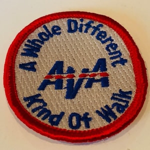 Pode incluir: Um remendo redondo branco com uma borda vermelha. O remendo tem um logotipo azul e vermelho com o texto "AAA" no centro. O texto "A Whole Different Kind Of Walk" circunda o logotipo.