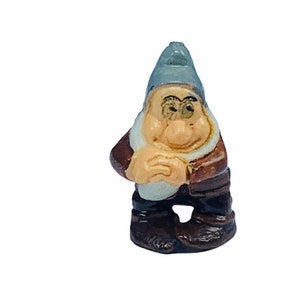 May include: A small ceramic figurine of a dwarf wearing a grey hat, brown jacket, and white collar. The dwarf has a brown beard and a smiling face.