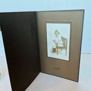 May include: A black and white photograph of a young girl sitting on a wooden bench. The girl is wearing a white dress and has her hair tied up with a ribbon. The photo is framed in a brown cardboard mount with a white border.