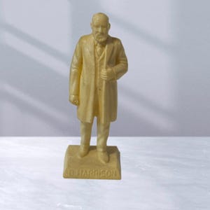 May include: A cream-colored plastic figurine of a man wearing a suit and coat. The figurine is standing on a base with the name "B. HARRISON" inscribed on it.