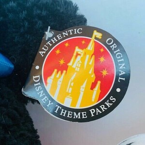 May include: A black plush toy with a red, yellow, and white tag that reads "Authentic Disney Theme Parks Original". The tag features a cartoon image of Cinderella's castle.