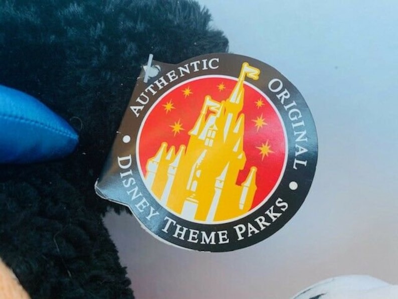 May include: A black plush toy with a red, yellow, and white tag that reads "Authentic Disney Theme Parks Original". The tag features a cartoon image of Cinderella's castle.
