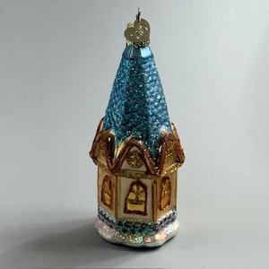 May include: Ornament shaped like a small church or building. The roof is a sparkling teal color, and the walls are a cream color with gold trim. The base has a glittery, iridescent appearance. A small gold heart hangs from the top.