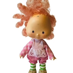 May include: A vintage Strawberry Shortcake doll with pink curly hair, wearing a pink and white gingham dress, pink trousers, green and white striped stockings, and brown shoes.