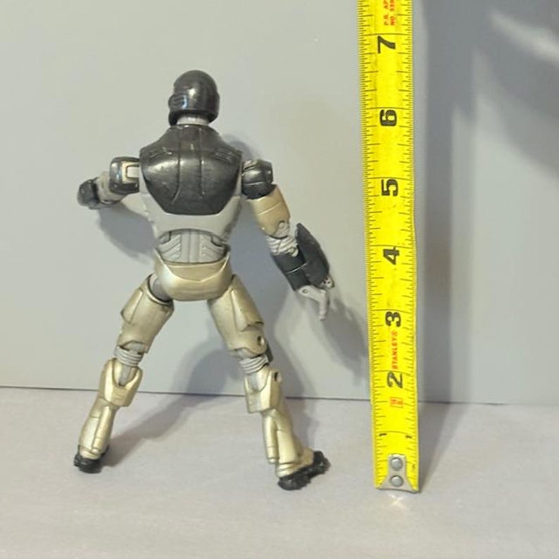 May include: A black and silver robot action figure standing approximately 6 inches tall. The figure has a black head and torso with silver arms and legs. The figure is posed with its arms raised and its legs spread apart.