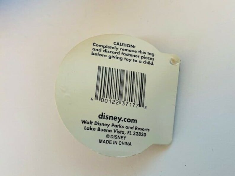 May include: White tag with black text and a barcode. The text reads "CAUTION: Completely remove this tag and discard fastener pieces before giving toy to a child."  The text also reads "disney.com Walt Disney Parks and Resorts Lake Buena Vista, FL 32830 &copy;DISNEY MADE IN CHINA"