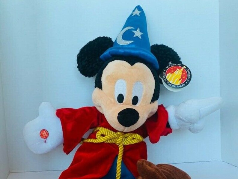 May include: A plush Mickey Mouse dressed as a wizard, wearing a blue hat with a crescent moon and star. He is wearing a red robe with a gold rope belt. The plush toy has a tag that says "Authentic Original Disney Theme Parks".