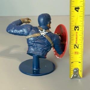 May include: A blue and red Captain America bust figurine with a raised arm holding a small object. The figurine is on a blue base and is holding a red, white and blue shield.