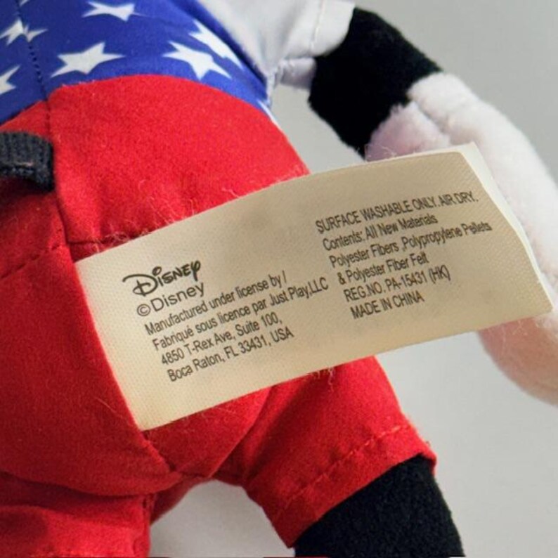 Op de afbeelding: Een close-up van een Disney pluche speelgoed. De kleding van het speelgoed heeft een rood-wit gestreept ontwerp, met een blauw gedeelte versierd met witte sterren. Een label met het Disney-logo en fabricagedetails is zichtbaar.