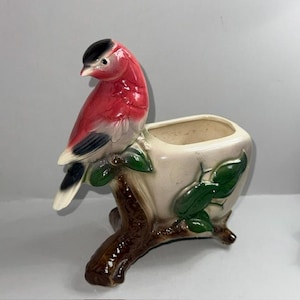 Pode incluir: Um vaso de cerâmica vintage em forma de pássaro empoleirado em um galho de árvore. O pássaro é vermelho, preto e branco. O vaso é bege com folhas verdes e um galho marrom.