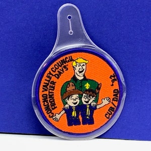 May include: A round patch with the text "Concho Valley Frontier Days" and "Cub/Dad '92" in a clear plastic holder. The patch features a cartoon of a man and two children in blue uniforms on an orange background.