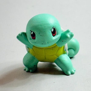 Puede incluir: Una peque&ntilde;a figura de Squirtle turquesa con caparaz&oacute;n amarillo. La figura tiene ojos rojos y est&aacute; posando con los brazos extendidos. Este juguete coleccionable es de la franquicia Pok&eacute;mon.