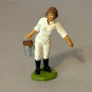 Puede incluir: Figura en miniatura de una mujer con mono blanco y botas negras, sosteniendo un cubo plateado lleno de material marrón. La figura está sobre una base verde, con un fondo neutro.